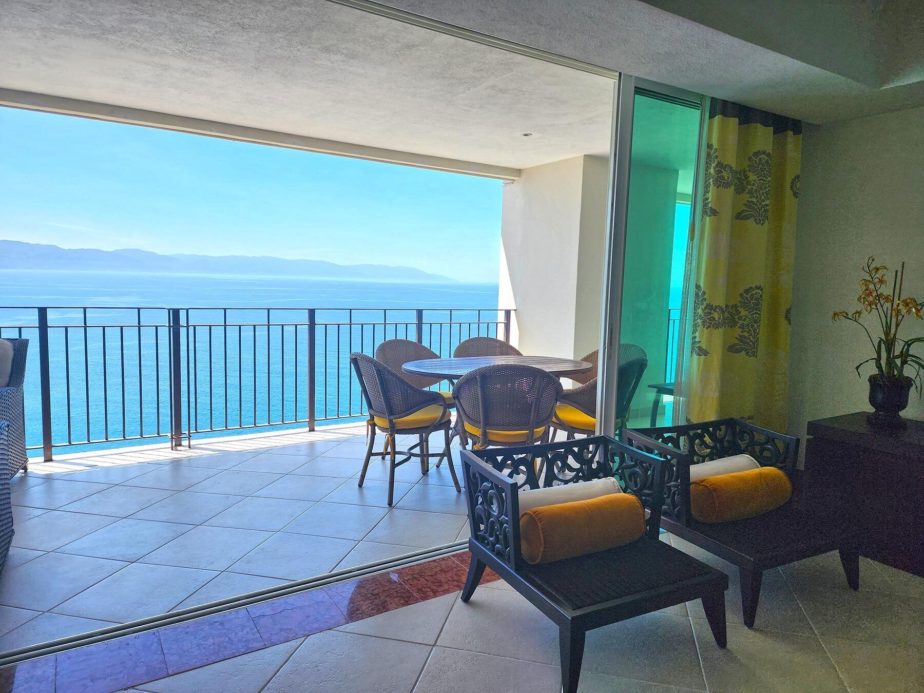 A balcony with a view of the sea, a table with chairs, and a sofa with cushions.