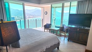 A bedroom with a large bed, a TV, and a balcony with a view of mountains and a city.