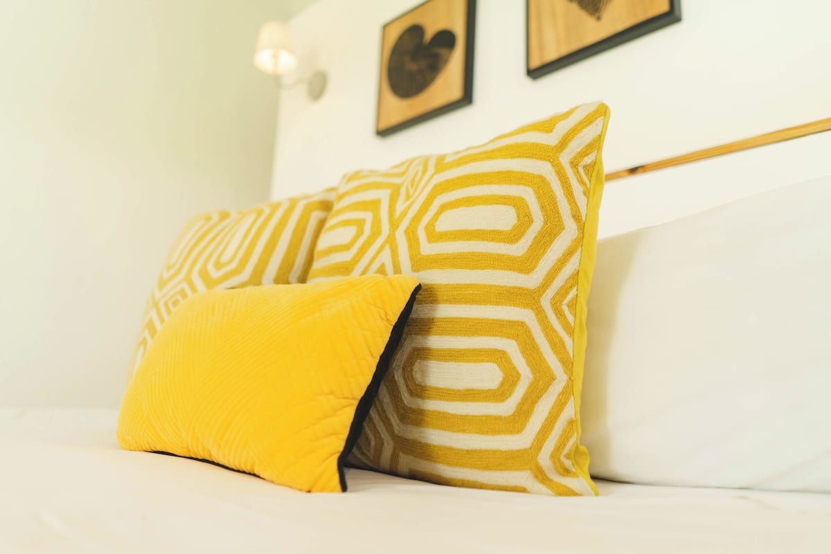 A bed with two pillows, one yellow and one with a geometric pattern, in a room with a white headboard and framed artwork on the wall.