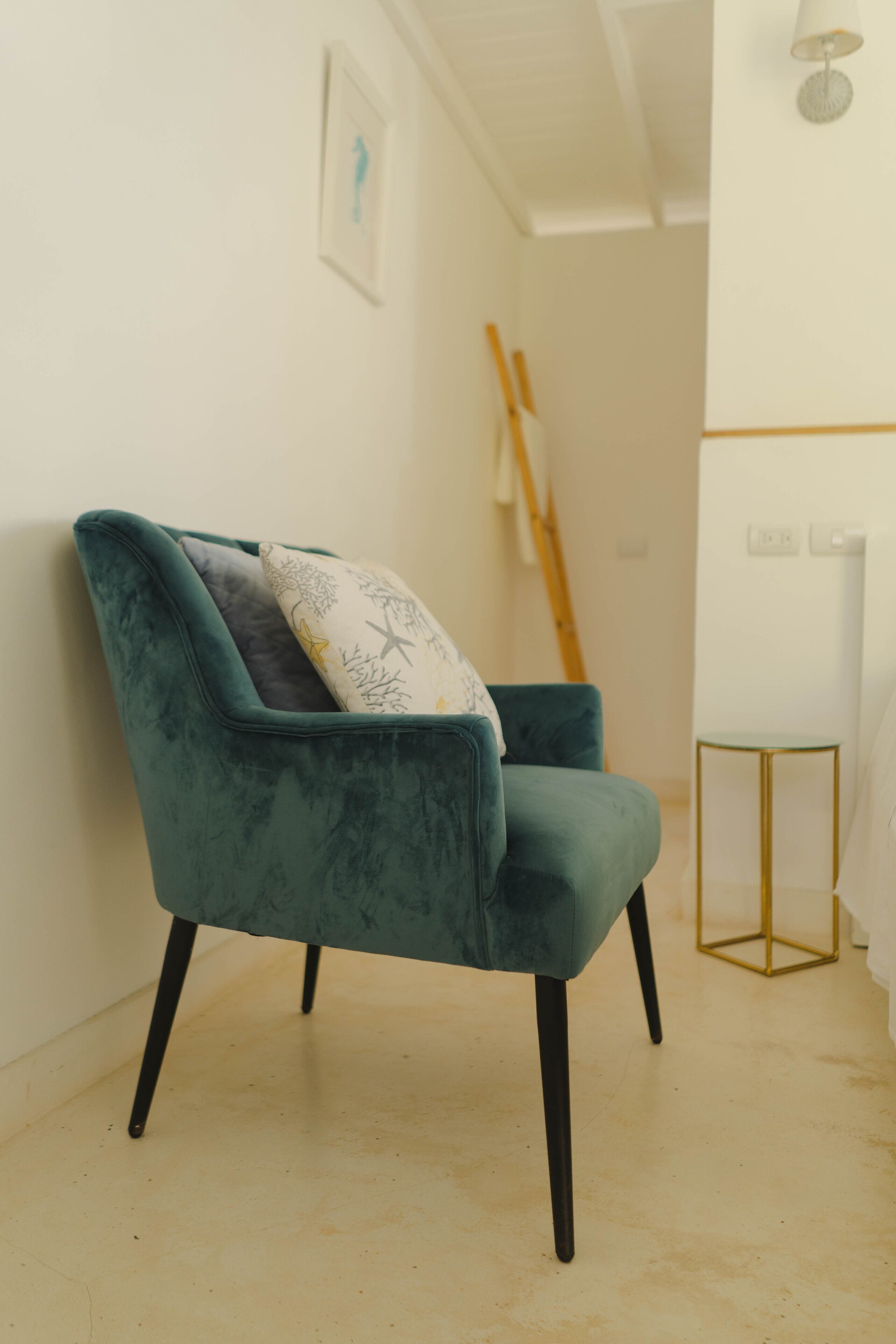 Un sillón tapizado en terciopelo azul verdoso con patas negras y un cojín estampado, ubicado en una habitación con suelo y paredes blancos, una pequeña mesa redonda con base dorada y una lámpara blanca en la pared.