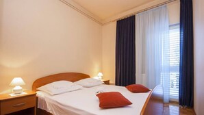 Una cama bien hecha con sábanas blancas y dos almohadas rojas, un cabecero de madera y una mesita de noche con una lámpara.
