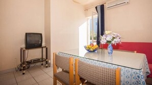 Un pequeño comedor con una mesa de vidrio, dos sillas, un frutero con fruta, un jarrón con flores, un televisor sobre un mueble bajo y un sofá rojo.