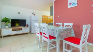 A dining area with a table set for four, white chairs with red cushions, a television on a wooden cabinet, and a kitchen in the background. - Apartments Alaga - B4+2 (Tribunj)