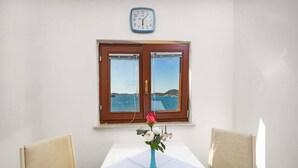 Una habitación con una mesa blanca, dos sillas, un jarrón azul con flores, un reloj y una ventana con vista al mar.