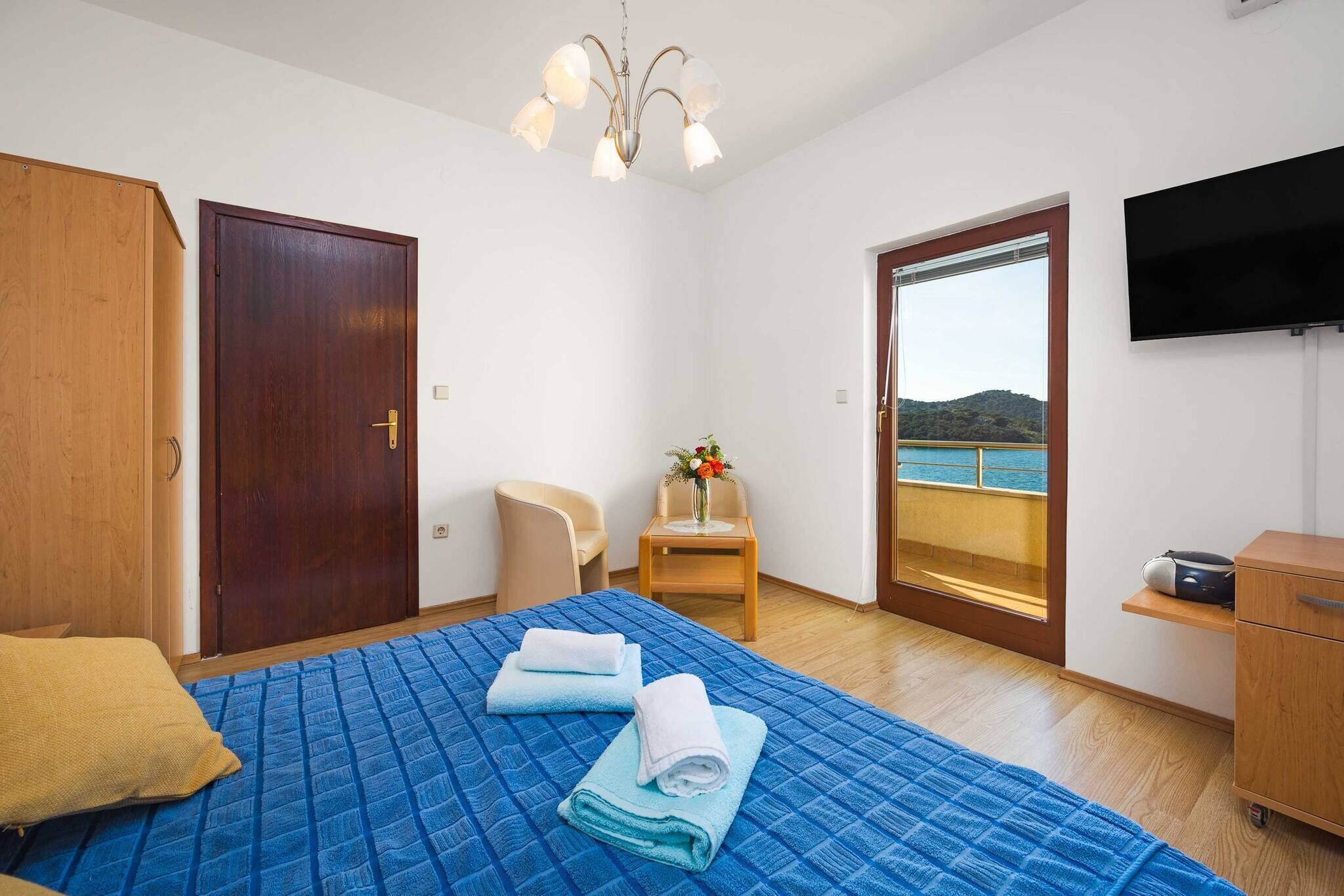 A hotel room with a bed, a chair, a TV, a wooden wardrobe, and a door leading to a balcony with a view of the sea.