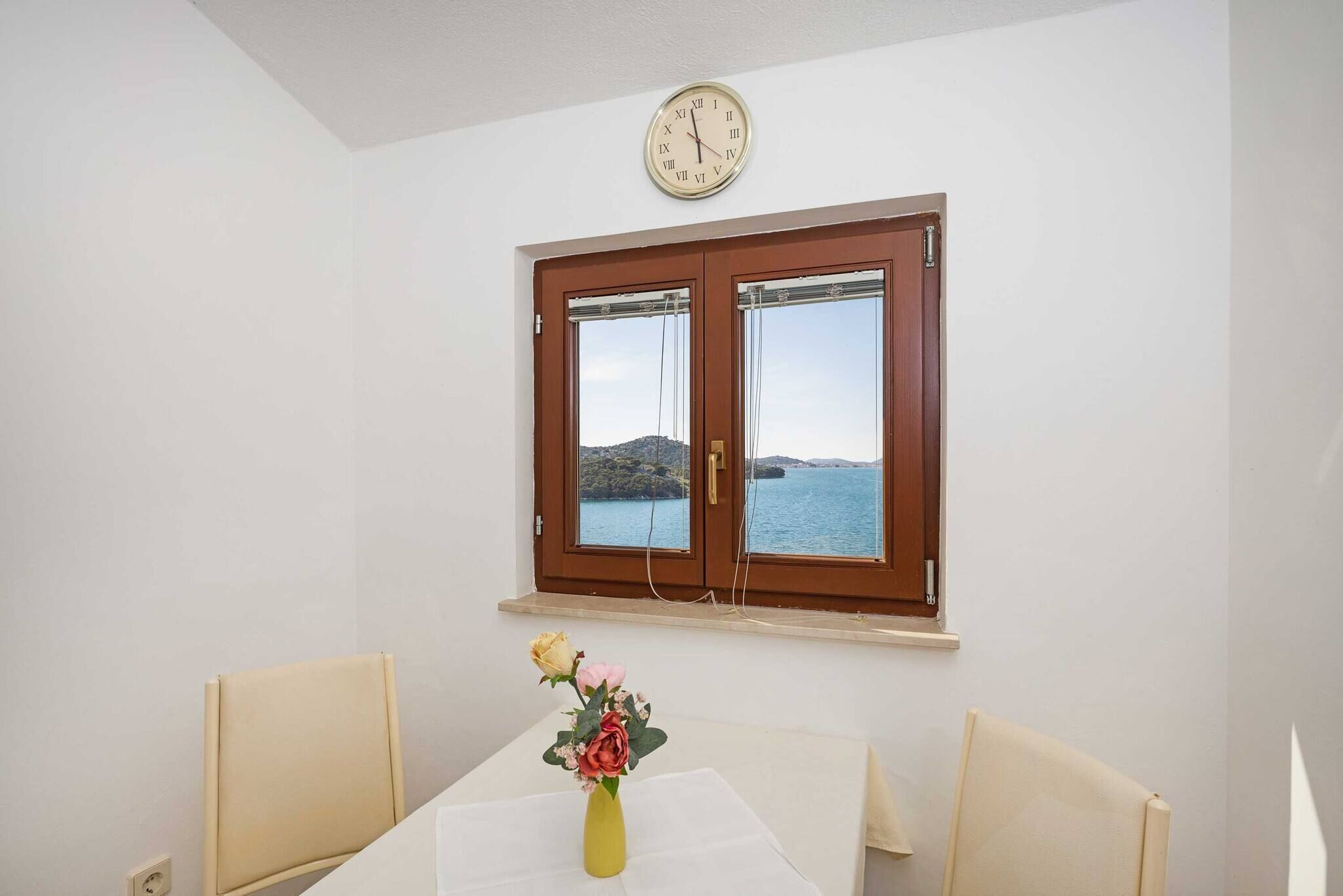 Een kamer met een tafel, twee stoelen, een vaas met bloemen, een klok en een raam met uitzicht op zee.