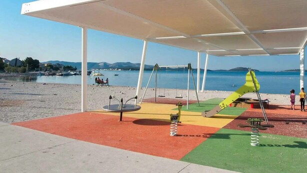 A covered outdoor area with a playground, including swings and a slide, near a body of water. - Apartments Maretic Brodarica A1 (Šibenik-Brodarica)