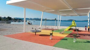 A covered outdoor area with a playground, including swings and a slide, near a body of water. - Apartments Maretic Brodarica A2 (Šibenik-Brodarica)