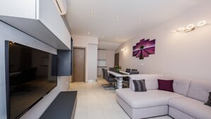 Ein modernes Wohnzimmer mit großem Flachbildfernseher, einem Ecksofa, einem Essbereich mit Tisch und Stühlen sowie einer Küche im Hintergrund.