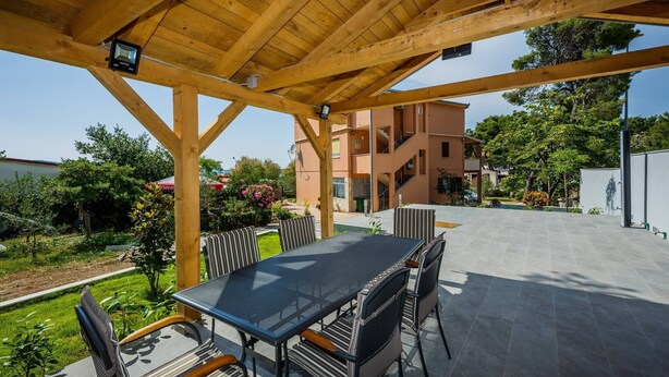 An outdoor patio with a wooden pergola, a table with chairs, and a view of a building in the background. - Beachfront One Bedroom Apartment Savin (Šibenik-Brodarica)