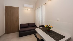 A modern, compact living space with a wooden door, a dark sofa, a dining table with a vase of flowers, and a wall-mounted air conditioner. - Beachfront One Bedroom Apartment Savin (Šibenik-Brodarica)