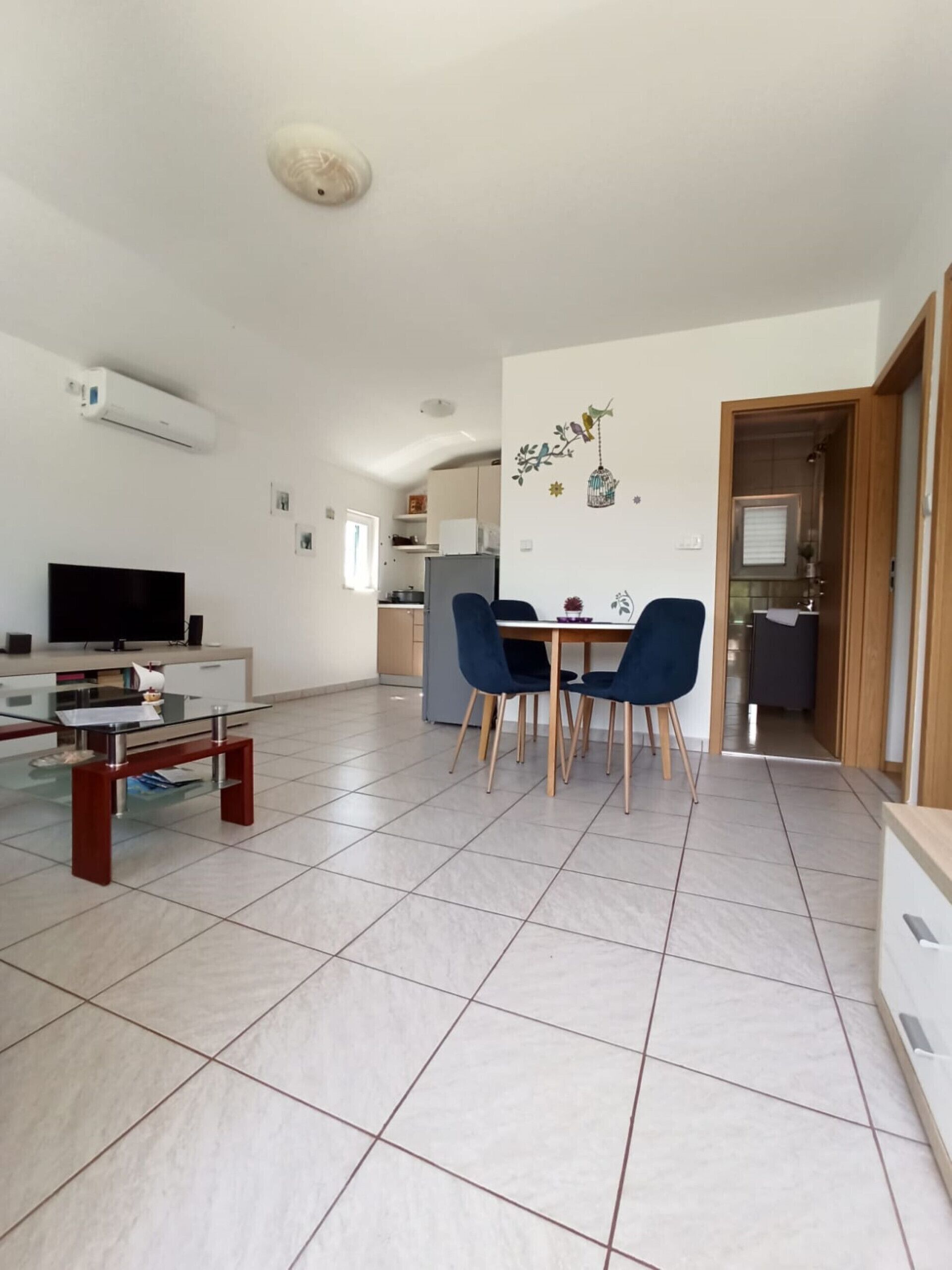 A modern living room with a glass coffee table, a flat-screen TV, and a dining area with blue chairs.