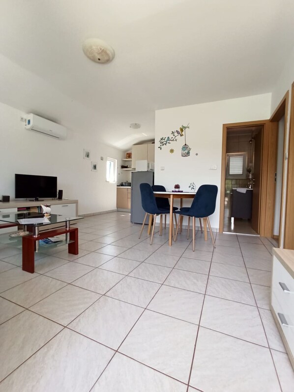 A modern living room with a glass coffee table, a flat-screen TV, and a dining area with blue chairs.