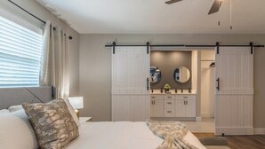 Une chambre avec un lit, une grande fenêtre avec des stores, un ventilateur de plafond et une porte coulissante de style rustique donnant sur une salle de bain.