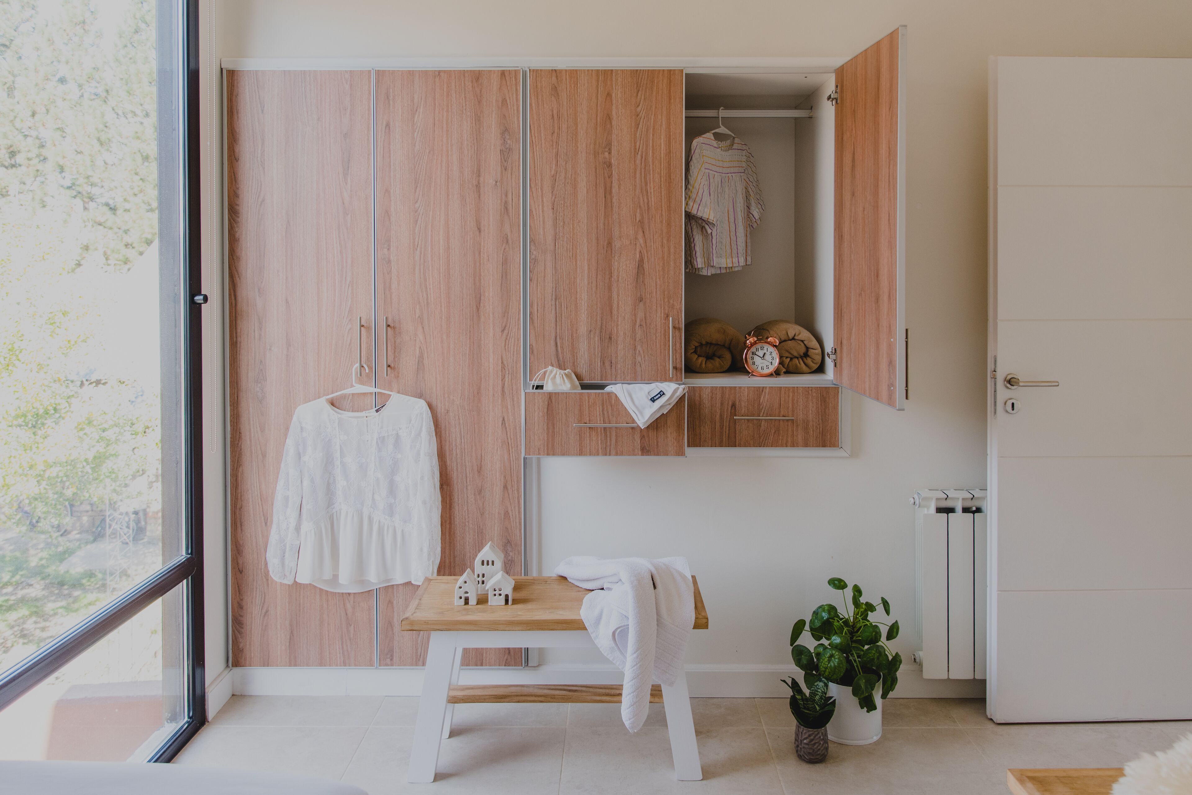 Een slaapkamer met een houten garderobe, een klein tafeltje met een witte handdoek, een hangende kledingstuk en een kamerplant.