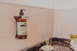 Un baño con una botella de jabón JABON U en un jabonera de pared, un vaso de agua y una pequeña planta en maceta sobre el mostrador.