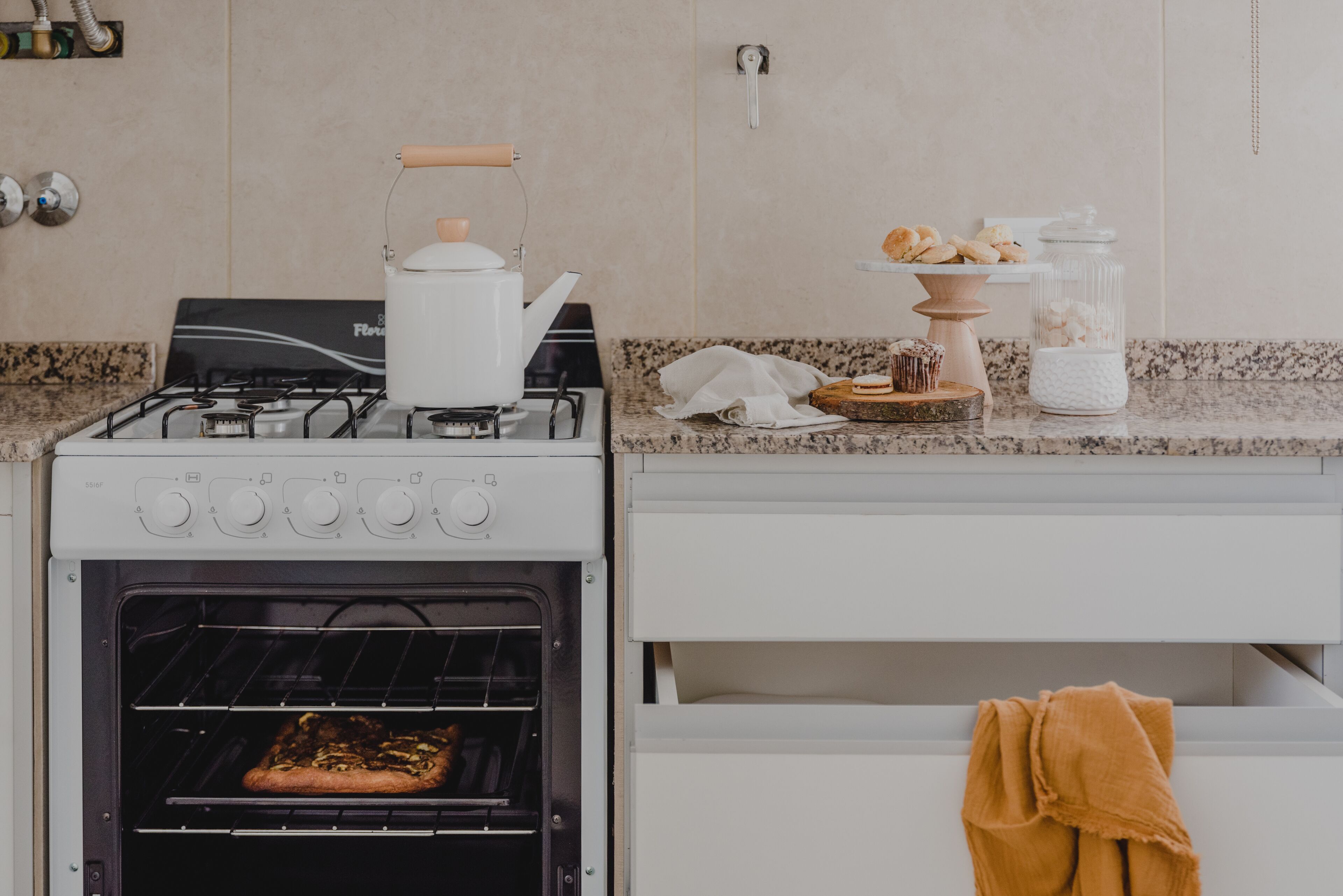 Een keuken met een gasfornuis, een oven, een koffiezetapparaat, een broodvitrine en een suikerpot.