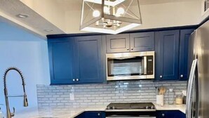 Een moderne keuken met blauwe kastjes, een roestvrijstalen koelkast, een inbouwoven en een bijzondere geometrische hanglamp.