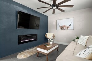 Un salon moderne doté d’une cheminée, d’un ventilateur de plafond, d’un téléviseur fixé au mur et d’un canapé agrémenté de coussins décoratifs.