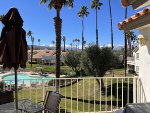 A balcony with a view of a swimming pool, palm trees, and a mountain range in the background. - Cozy Desert Escape Available! 2BR Condo Golf & Pool (Palm Desert)