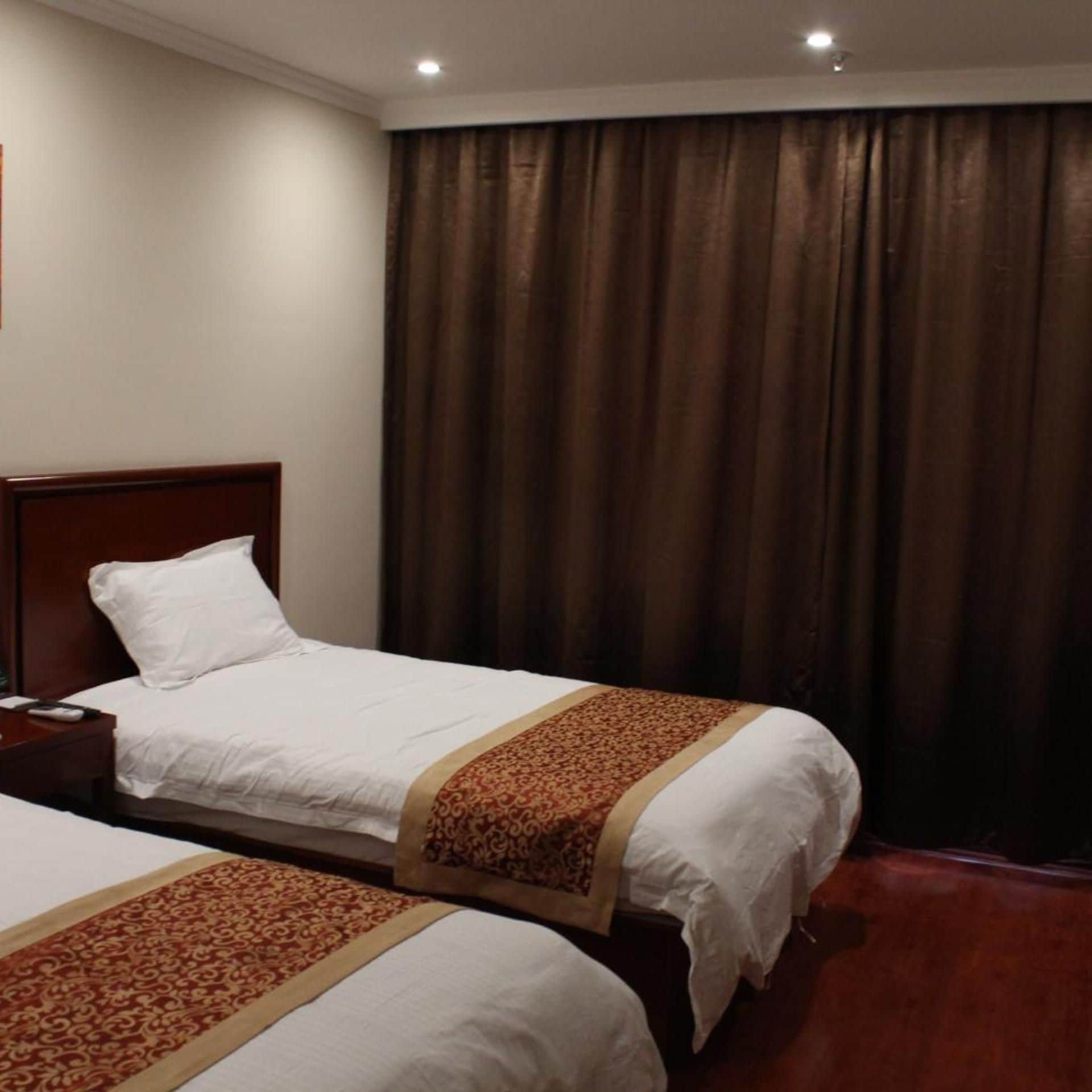 Ein Hotelzimmer mit zwei Einzelbetten, einem hölzernen Kopfteil, einem Nachttisch mit Lampe und einem großen Fenster mit Vorhängen.