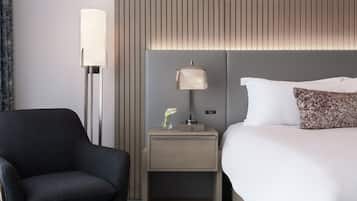 Habitación de hotel con cama, mesita de noche con lámpara, una silla y un muro con persianas verticales.