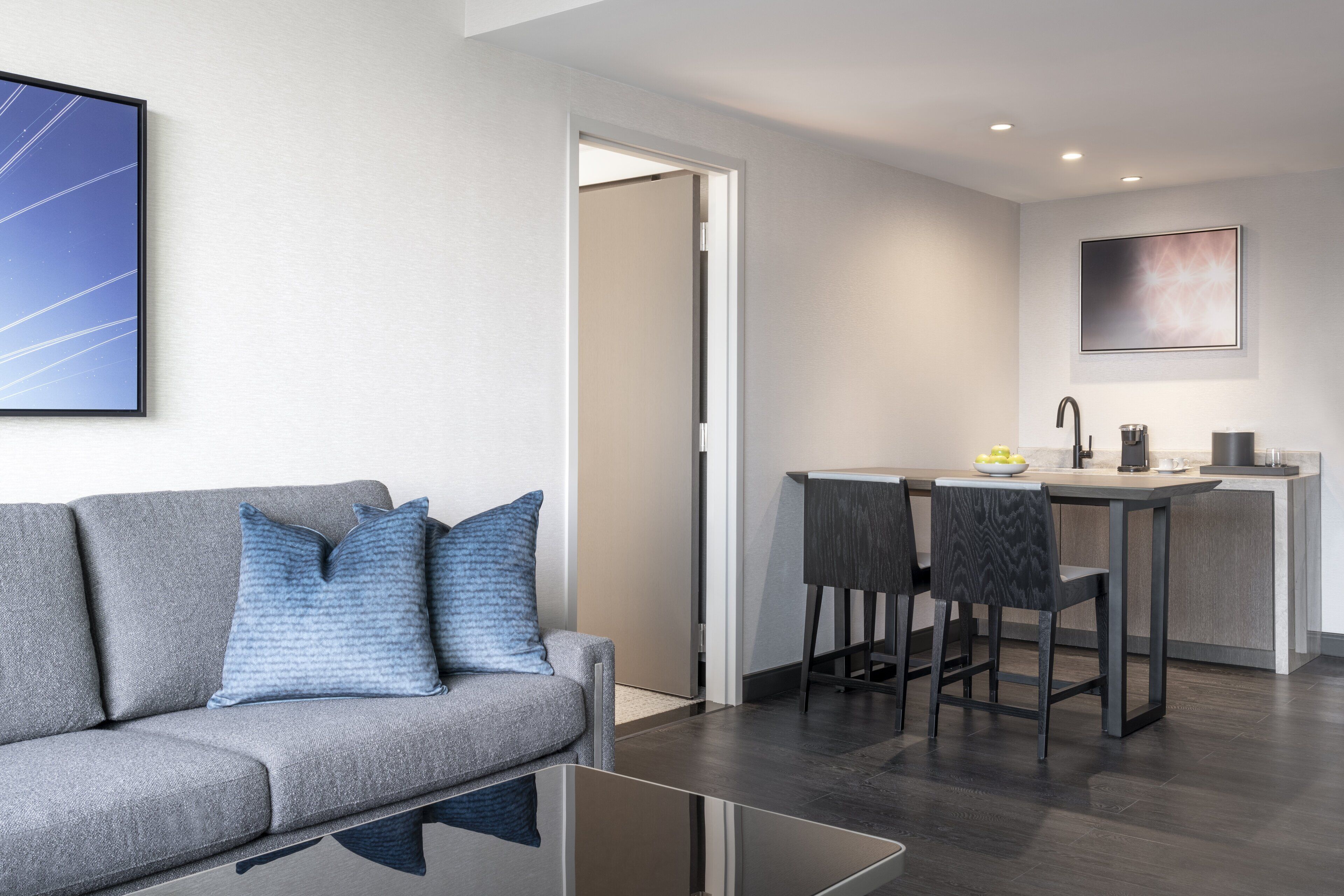 A modern living room with a grey sofa, two blue pillows, and a glass coffee table. A kitchen area with a bar stool and a small island is visible in the background.