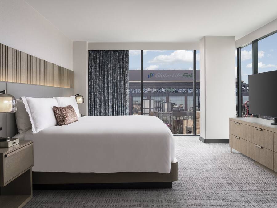 Quarto de hotel moderno com uma cama grande, uma televisão, uma escrivaninha com luminária e vista para a cidade através de amplas janelas.