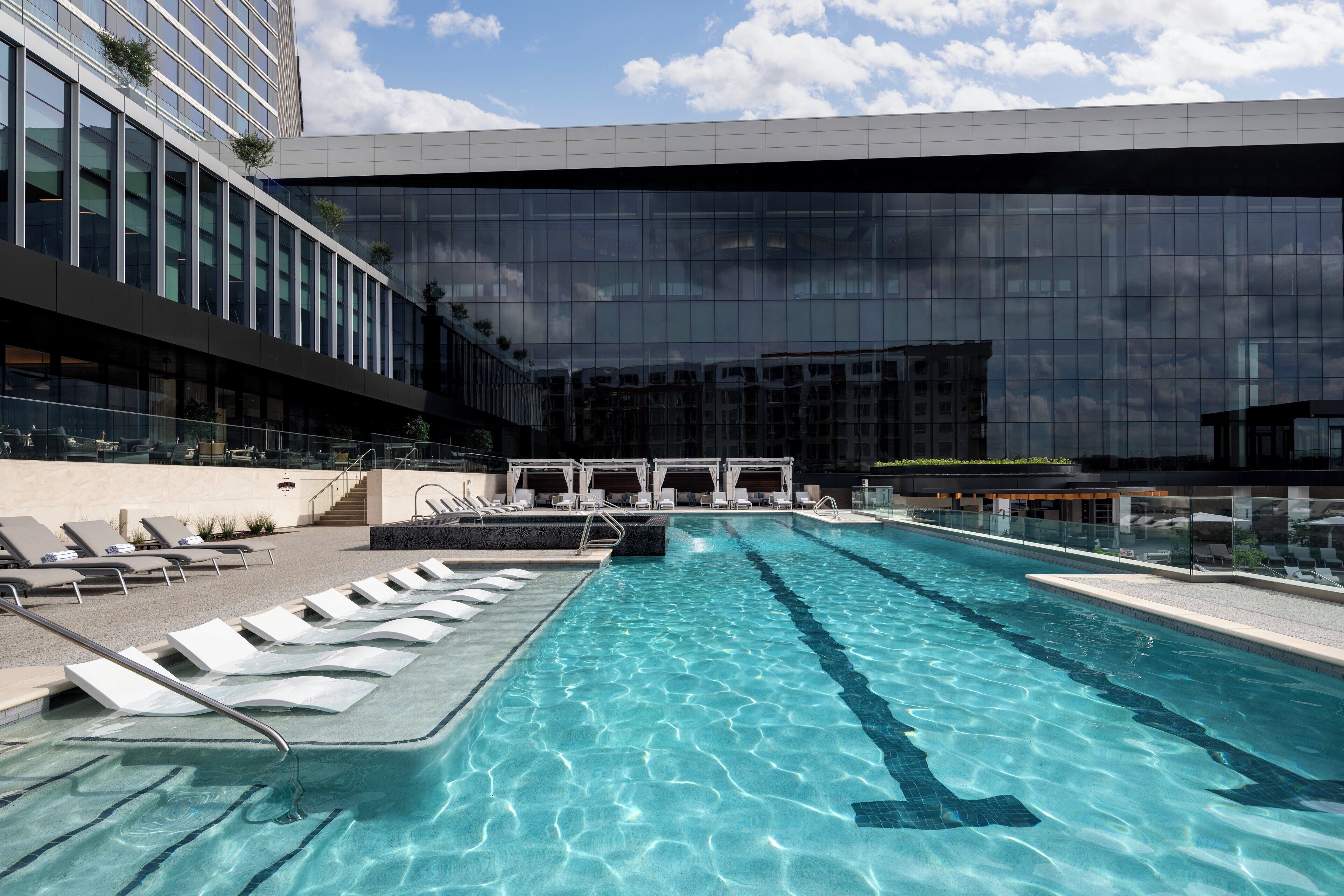 A modern hotel with a large indoor swimming pool, lounge chairs, and a glass facade reflecting the sky.