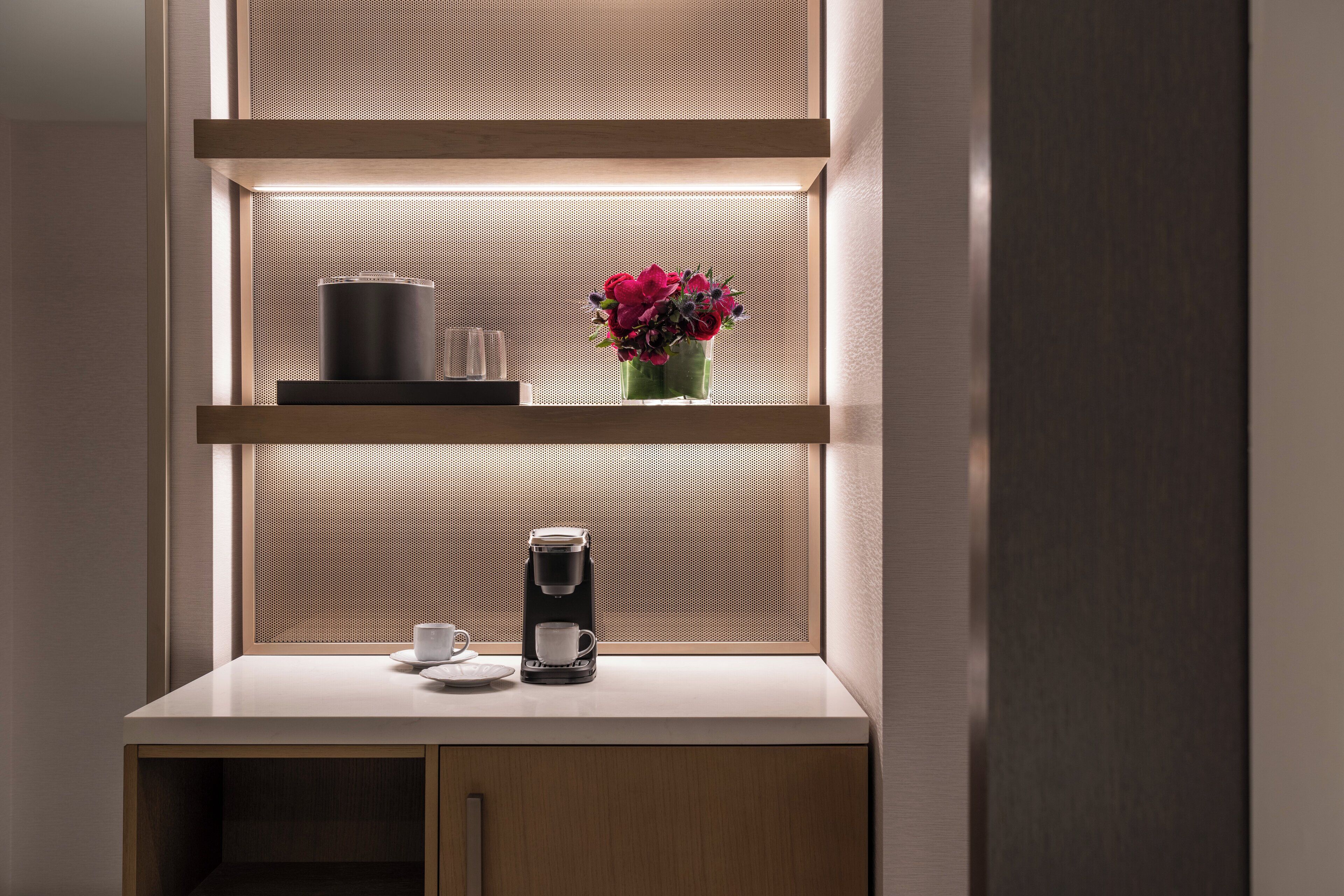 Una kitchenette moderna con una cafetera, una taza y un jarrón de flores sobre una repisa.