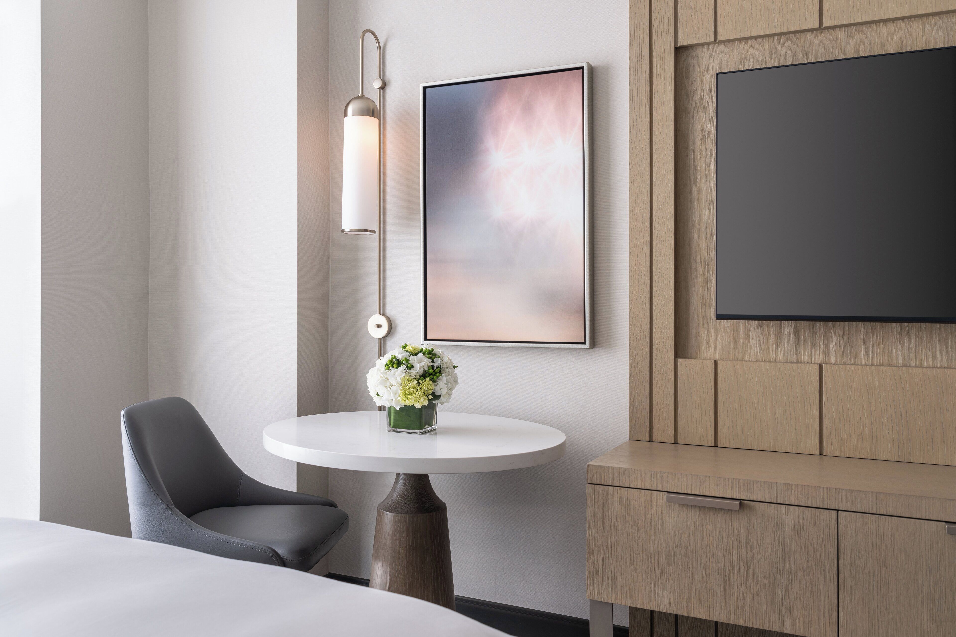 A modern hotel room with a bed, a round table with a vase of flowers, a gray chair, a wall-mounted light fixture, and a framed abstract painting.