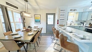 Un espace repas moderne avec une longue table dressée pour six personnes, une cuisine dotée de meubles blancs et une œuvre d’art encadrée accrochée au mur.