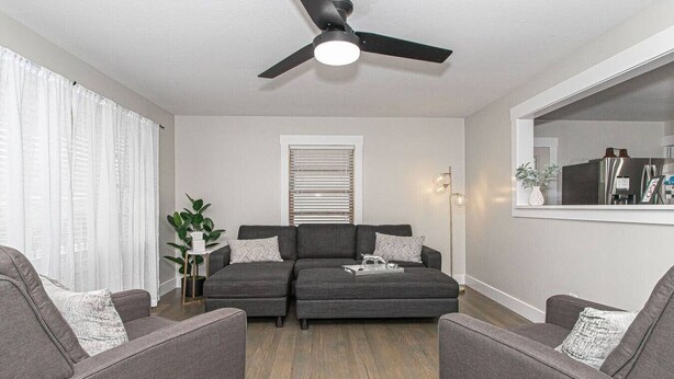 Una sala moderna con un sofá gris, dos sillones, un ventilador de techo y un espejo grande que refleja una cocina.