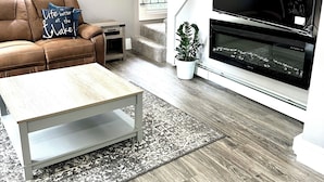 Un salon moderne avec un canapé en cuir marron, une table basse en bois, un téléviseur noir posé sur un meuble blanc et un escalier menant à un autre niveau.