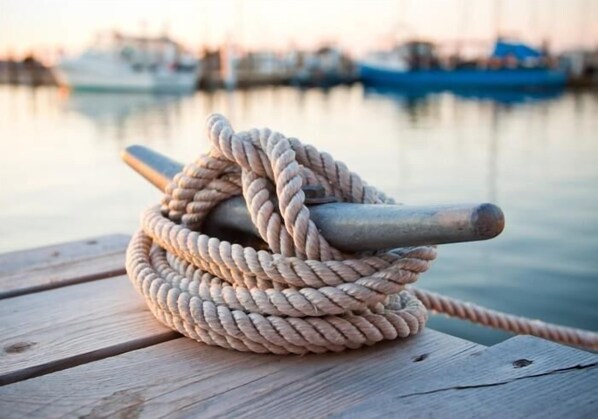 Cuerda enrollada sobre una superficie de madera, con un objeto metálico, probablemente un pasamanos, y un cuerpo de agua con barcos al fondo.