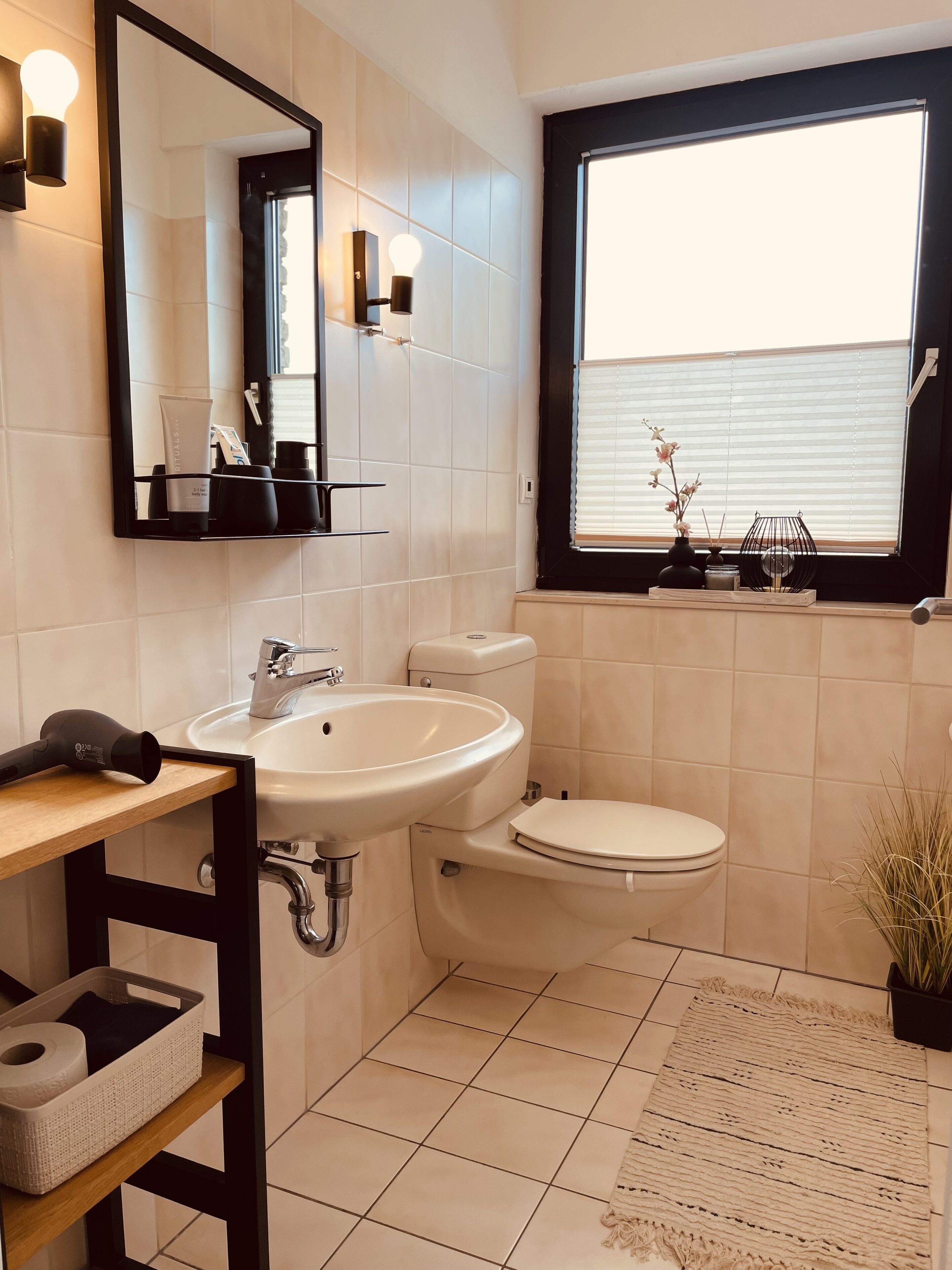 Ein Badezimmer mit einem weißen Waschbecken, einer Toilette, einem Spiegel und einem Fenster mit Jalousien.