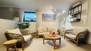 Un salon avec des meubles en osier, une table basse avec des collations, un radiateur, une fenêtre avec des rideaux et un tableau représentant une plage accroché au mur.