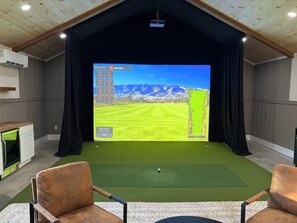 Une pièce dotée d’un grand écran diffusant une simulation de golf, de deux fauteuils en cuir, d’une petite table et d’un projecteur fixé au plafond.