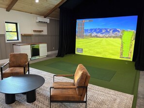 Una habitación con una pantalla grande mostrando un campo de golf, dos sillones de cuero, una mesita, una zona de cocina y una ventana.