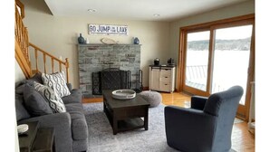Un salon doté d’une cheminée en pierre, d’un canapé, d’un fauteuil et offrant une vue sur un lac grâce à de grandes fenêtres.
