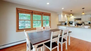 Un coin repas avec une table en bois et des chaises blanches, une grande fenêtre donnant sur un paysage verdoyant, et une cuisine avec des meubles blancs et des appareils électroménagers modernes.