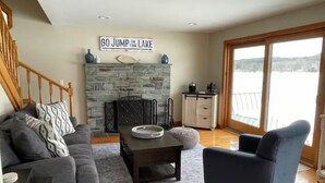Una sala de estar con chimenea de piedra, un sofá, un sillón y vistas a un lago a través de amplios ventanales.