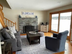 Ein Wohnzimmer mit Kamin aus Stein, einer Couch, einem Sessel und Blick auf einen See durch große Fenster.