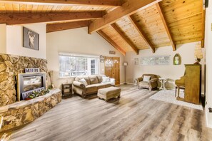 A spacious living room with a stone fireplace, wooden beams, a large window, and a piano.