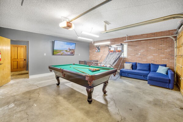 A pool table in a room with a blue couch, a painting on the wall, and a brick wall.