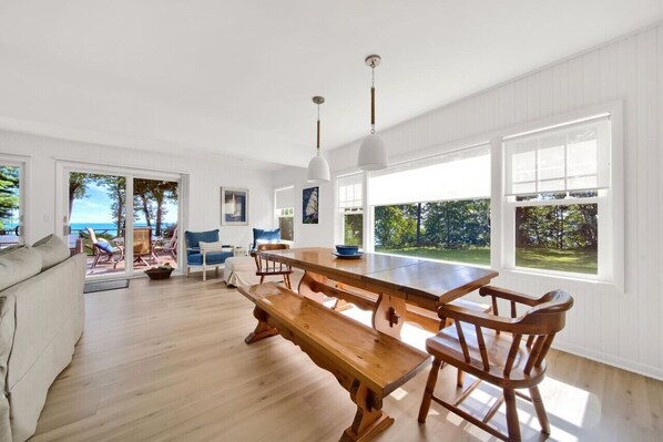 A spacious dining area with a long wooden table and benches, large windows with a view of greenery, and a white interior with wooden flooring.