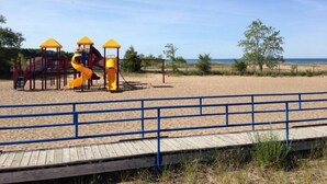 Ein Strandbereich mit Sand, einem Spielplatz mit einer gelben Rutsche und einem hölzernen Steg im Vordergrund.