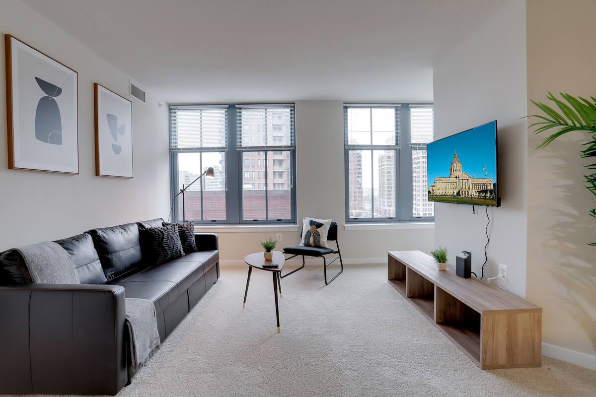 Ein modernes Wohnzimmer mit einem schwarzen Ledersofa, einem Holztisch, einem schwarzen Sessel und einem an der Wand befestigten Fernseher.