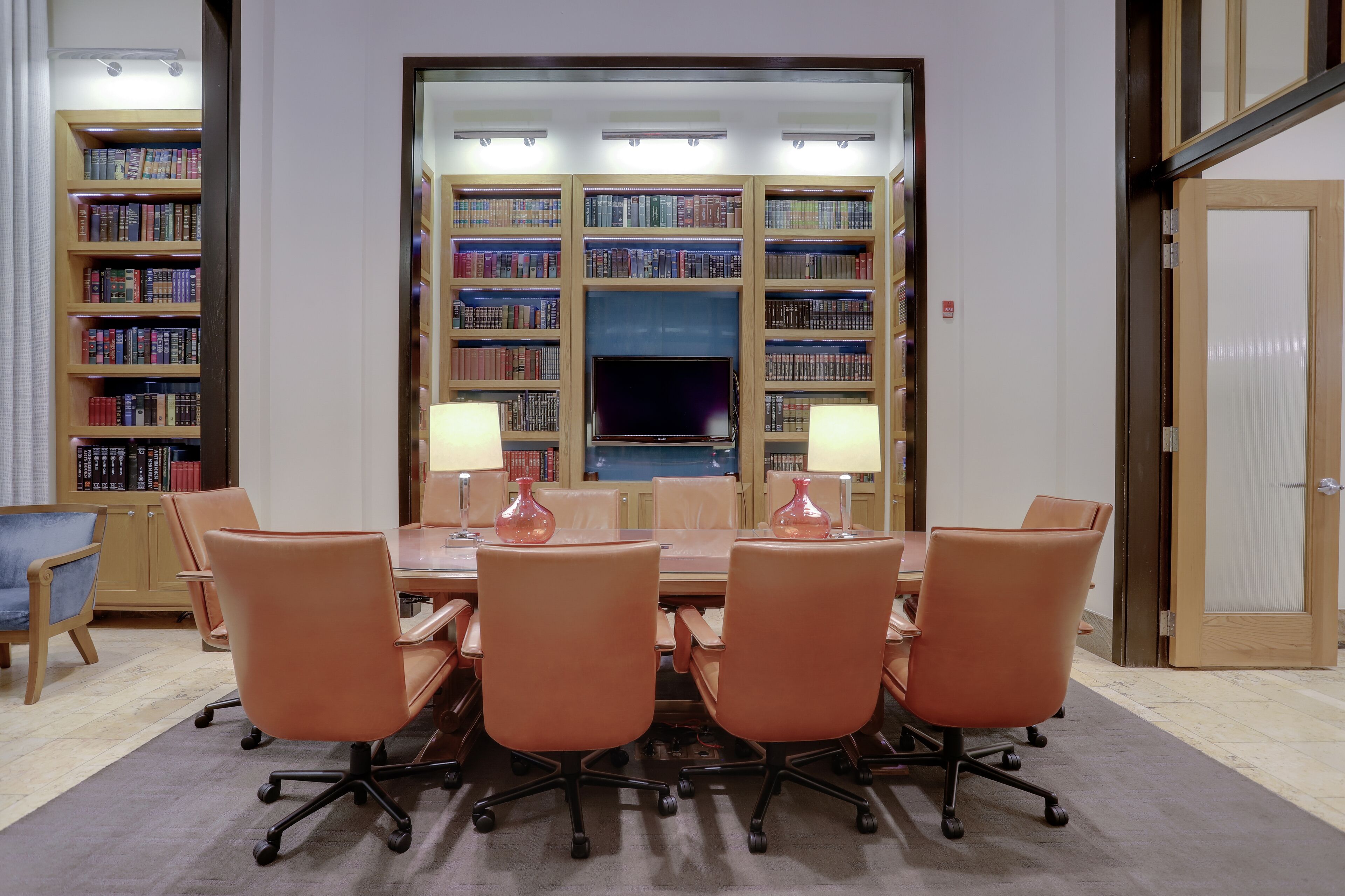 A conference room with a long table, six chairs, two lamps, and a bookshelf in the background.