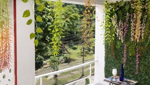 A balcony with hanging plants, a small table with a vase, and a view of a garden. - Breathe in a quiet and safe forest  (Cali)